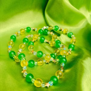 Puede incluir: Tres pulseras de cuentas con cuentas verdes y amarillas y detalles de estrellas doradas descansan sobre una vibrante tela de satén verde. Las pulseras presentan una mezcla de cuentas redondas y facetadas, creando un aspecto en capas y texturizado. La estética general es brillante y juguetona.
