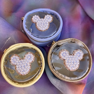 May include: Three round, clear cosmetic bags with a Mickey Mouse design. Each bag features a pearl-studded Mickey Mouse head, a gold-coloured trim, and a zip closure. The bags are in pastel yellow, blue, and gold.