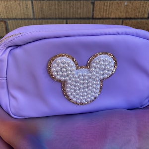 Puede incluir: Un neceser lavanda con cremallera dorada y una cabeza decorativa de Mickey Mouse hecha de perlas blancas y purpurina dorada. La bolsa es rectangular y tiene forma redondeada.