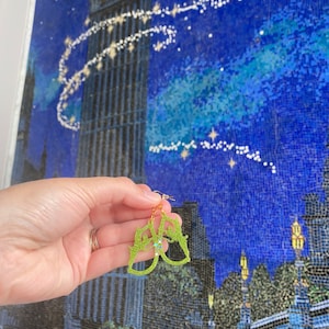 Puede incluir: Pendientes de purpurina verde en forma de silueta del castillo de Cenicienta con un acento de purpurina azul. Los pendientes se muestran sobre un fondo de mosaico de Big Ben y un cielo nocturno.