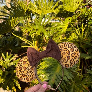 May include: Brown headband with a brown bow and two leopard print ears. The headband is held in a hand.