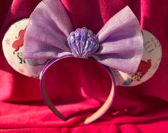 May include: A purple headband with mouse ears. The ears are white with a cartoon image of a mermaid and the text "AS THE SEA". A large purple bow and a seashell embellishment are centered on the headband.