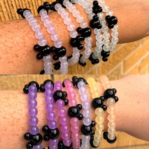 May include: A set of six colorful beaded bracelets with black Mickey Mouse head charms. The bracelets are made of translucent beads in shades of purple, pink, yellow, and white.