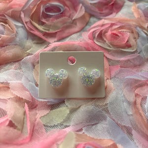 May include: A pair of white glitter Mickey Mouse stud earrings on a white card.