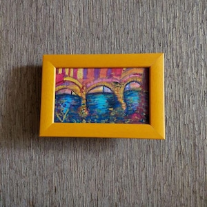 K&ouml;nnte beinhalten: Ein kleines, gerahmtes Gem&auml;lde mit einer abstrakten Darstellung von B&ouml;gen &uuml;ber Wasser. Das Kunstwerk zeigt eine Palette aus Blau-, Rosa- und Gelbt&ouml;nen, eingefasst in einen leuchtend gelben Rahmen. Das Gem&auml;lde wird vor einer strukturierten, neutral get&ouml;nten Wand pr&auml;sentiert.