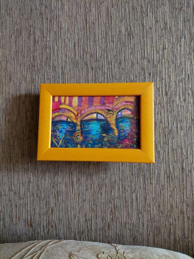 K&ouml;nnte beinhalten: Ein kleines, gerahmtes Gem&auml;lde mit einer abstrakten Darstellung von B&ouml;gen &uuml;ber Wasser. Das Kunstwerk zeigt eine Palette aus Blau-, Rosa- und Gelbt&ouml;nen, eingefasst in einen leuchtend gelben Rahmen. Das Gem&auml;lde wird vor einer strukturierten, neutral get&ouml;nten Wand pr&auml;sentiert.