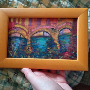 K&ouml;nnte beinhalten: Ein gerahmtes abstraktes Gem&auml;lde in einem hellen Holzrahmen. Das Kunstwerk zeigt drei bogenf&ouml;rmige Strukturen in Rosa, Orange und Lila, &uuml;ber einer blau-gr&uuml;nen Wasserszene. Das Gem&auml;lde wird von einer Hand gehalten.