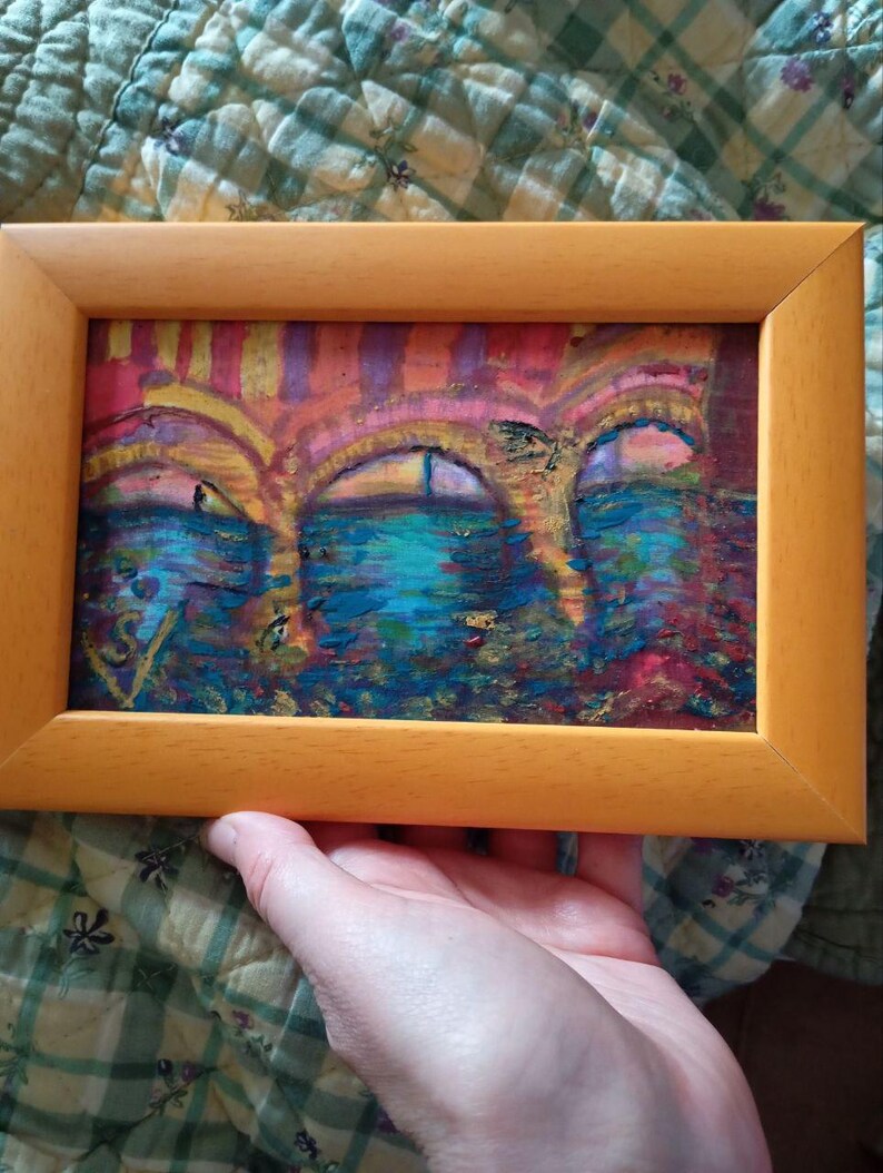 K&ouml;nnte beinhalten: Ein gerahmtes abstraktes Gem&auml;lde in einem hellen Holzrahmen. Das Kunstwerk zeigt drei bogenf&ouml;rmige Strukturen in Rosa, Orange und Lila, &uuml;ber einer blau-gr&uuml;nen Wasserszene. Das Gem&auml;lde wird von einer Hand gehalten.