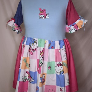 May include: A blue and pink dress with a patchwork skirt featuring Hello Kitty and her friends. The dress has short sleeves and a round neckline. The skirt is made of a variety of fabrics and colours, including pink, blue, green, and white.