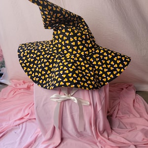 May include: A black witch's hat patterned with candy corn. The hat features a broad brim and a tall, pointed crown. A white ribbon is tied around the base.
