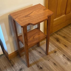 May include: A small, two-tiered wooden side table with a natural finish. The table has a rectangular top with a cutout on one side and a lower shelf. The legs are straight and the table is made of wood.