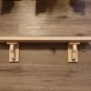 May include: A wooden shelf with two decorative brackets. The shelf is made of light wood and the brackets are a slightly darker shade of wood.