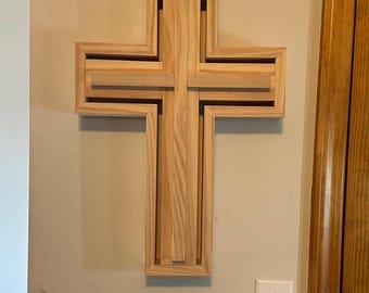May include: A wall-mounted wooden cross featuring a layered, three-dimensional design. The structure is composed of light-colored natural wood with visible grain, consisting of a central vertical beam and a horizontal crossbar set within a frame.