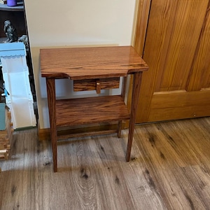 May include: A rectangular wooden side table with a polished brown finish and visible wood grain. The table features a flat top surface, a single small central drawer with a vertical handle, and a lower open shelf supported by four straight legs.