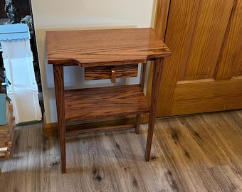 May include: A rectangular wooden side table with a polished brown finish and visible wood grain. The table features a flat top surface, a single small central drawer with a vertical handle, and a lower open shelf supported by four straight legs.