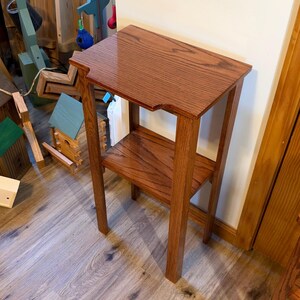 May include: A tall, narrow wooden side table with a rich, reddish-brown stained finish and a visible wood grain. The table features a rectangular top with notched corners and a single lower shelf supported by four straight, vertical legs.