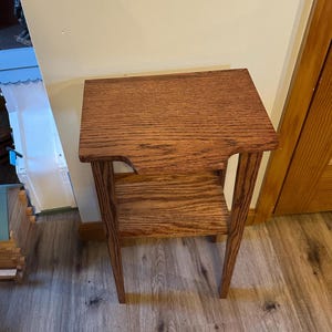 May include: A small wooden side table featuring a rectangular top with a notched front edge and a lower shelf. The piece is constructed from medium-brown wood with a visible grain pattern, supported by four straight legs in a simple design.