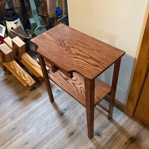May include: A small, wooden side table with a dark brown finish. The table has a rectangular top with a decorative edge and a lower shelf. The table is approximately 30 inches tall and is supported by four slender legs.