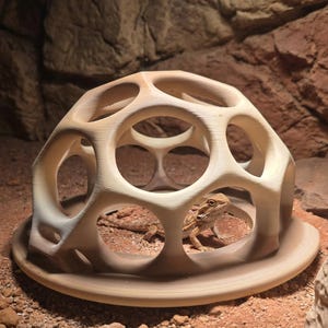 May include: A beige, dome-shaped reptile hide with a geometric, multi-holed lattice structure. The structure features a circular base and a smooth, matte finish. A small, light brown bearded dragon is positioned inside the hollow center of the dome.