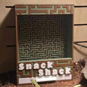 May include: A rectangular brown and light green box with a maze pattern on the back panel and the text "snack shack" in white letters on the front. The structure features an open front design and is positioned within a terrarium environment.