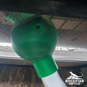 May include: A green, 3D-printed funnel-shaped attachment secured to a metal mesh screen. The object features a wide, rounded top section with a small circular opening and a narrower cylindrical base connected to a white tube. Includes Rockstar Reptiles logo.