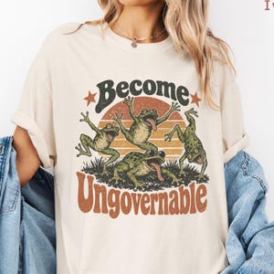 Peut inclure: Un t-shirt couleur crème arborant une impression graphique de trois grenouilles vertes dans des poses dynamiques sur un fond de coucher de soleil. Le texte "Become Ungovernable" est imprimé dans une police rétro sous les grenouilles, avec deux étoiles.