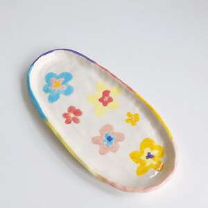 Può includere: Un vassoio ovale in ceramica bianca con un bordo arcobaleno e fiori dipinti in tonalità di blu, giallo, rosa e rosso.