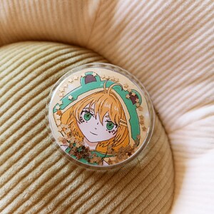 Op de afbeelding: Een ronde, heldere acrylspeld met een anime-stijl illustratie van een personage met groene ogen en oranje haar, die een groene kikkerhoed draagt. De speld is gevuld met gouden stervormige glitter, tegen een lichtbeige achtergrond.