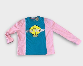 Può includere: Una camicia a maniche lunghe con un design a blocchi di colore. La camicia presenta un pannello centrale turchese con una grafica di cervello giallo e le parole "USE YOUR". Le maniche sono rosa.