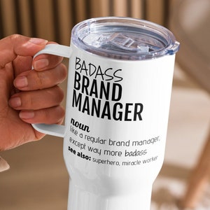 May include: White travel mug with a handle and a clear lid. The mug features the text "BADASS BRAND MANAGER" in black, along with a definition. The mug is held by a person.