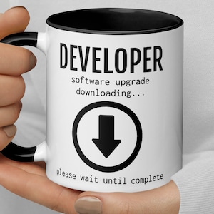 May include: White ceramic mug with a black rim and handle. The mug has black text that reads "DEVELOPER software upgrade downloading..." with a black downward arrow icon and the text "please wait until complete".