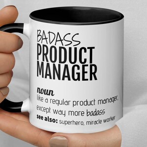 Peut inclure: Mug en céramique blanche avec intérieur et anse noirs. Le mug affiche le texte "BADASS PRODUCT MANAGER" en gras noir, ainsi qu'une définition et des termes associés. Un cadeau parfait pour un chef de produit.