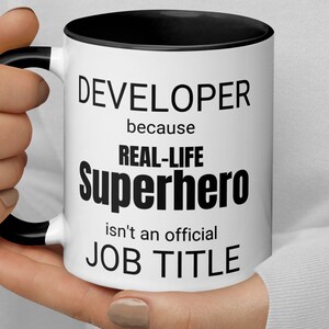 May include: White coffee mug with a black rim and handle. The mug has black text that reads "DEVELOPER because REAL-LIFE SUPERHERO isn't an official JOB TITLE".