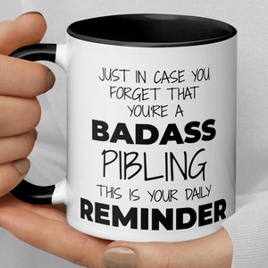 May include: White ceramic mug with a black rim and handle. The mug has black text that reads "Just in case you forget that you're a badass pibling. This is your daily reminder."