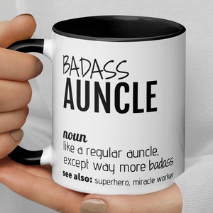 May include: White ceramic mug with a black rim and handle. The mug has black text that reads "BADASS AUNCLE" and a definition of the word "auncle" as "like a regular uncle, except way more badass." The text also says "see also: superhero, miracle worker."