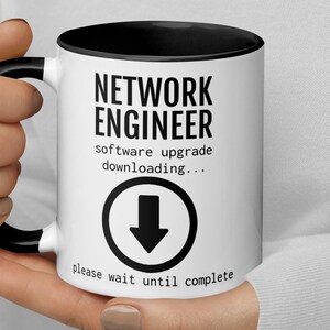 May include: White coffee mug with a black rim and handle. The mug has black text that reads "Network Engineer" and "software upgrade downloading..." with a downward arrow icon. The text at the bottom reads "please wait until complete."