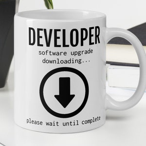 May include: White ceramic coffee mug with black text that reads "DEVELOPER software upgrade downloading..." with a black downward arrow icon in a circle and the text "please wait until complete".