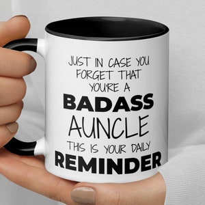 May include: White coffee mug with a black handle and black rim. The mug has black text that reads "Just in case you forget that you're a badass auncle. This is your daily reminder."