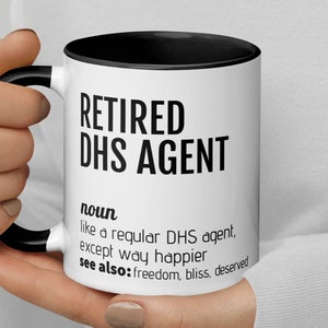 May include: White ceramic mug with a black interior and handle. The mug features the text "RETIRED DHS AGENT" in bold black letters, along with a definition and related terms. The mug is held by a person.