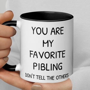 May include: White ceramic mug with a black rim and handle. The mug has black text that reads "You are my favourite sibling. Don't tell the others."