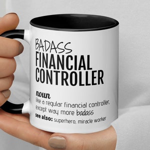 May include: White ceramic mug with a black rim and handle. The mug has black text that reads "BADASS FINANCIAL CONTROLLER noun like a regular financial controller. except way more badass see also: superhero, miracle worker"