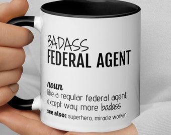May include: White coffee mug with a black rim and handle. The mug has black text that reads "BADASS FEDERAL AGENT" and a definition of the term.