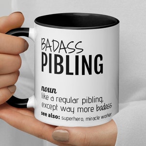 May include: White ceramic mug with a black rim and handle. The mug has black text that reads "BADASS PIBLING" and a definition of the word.