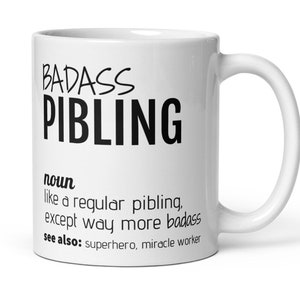 May include: White ceramic coffee mug with black text that reads "BADASS PIBLING" and a definition of the word. The definition reads "noun like a regular pibling, except way more badass see also: superhero, miracle worker"