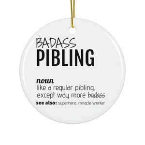 May include: A white, circular ceramic ornament featuring the words "BADASS PIBLING" in black. The definition below states "like a regular pibling, except way more badass." A gold string is attached for hanging.