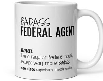 May include: White ceramic mug with the text "BADASS FEDERAL AGENT" in black. The mug includes a definition of the term, and the words "superhero, miracle worker" are also included.