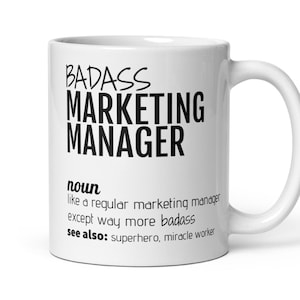 May include: White ceramic mug with the text "BADASS MARKETING MANAGER" in black, along with a definition and related terms. The mug has a curved handle and is designed for hot beverages.
