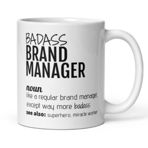 May include: White ceramic coffee mug with black text that reads "BADASS BRAND MANAGER noun like a regular brand manager, except way more badass see also: superhero, miracle worker"