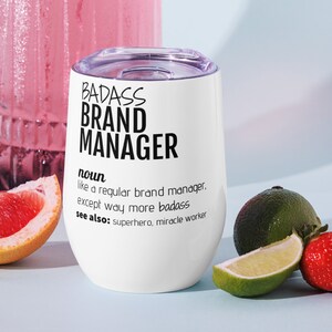 May include: White stainless steel wine tumbler with a black inscription that reads "BADASS BRAND MANAGER noun like a regular brand manager, except way more badass see also: superhero, miracle worker"