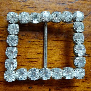 Puede incluir: Hebilla rectangular plateada con una fila de brillantes pedrería transparente y brillante alrededor del perímetro. Una barra plateada está centrada dentro del rectángulo. La hebilla está sobre una superficie de madera cálida.